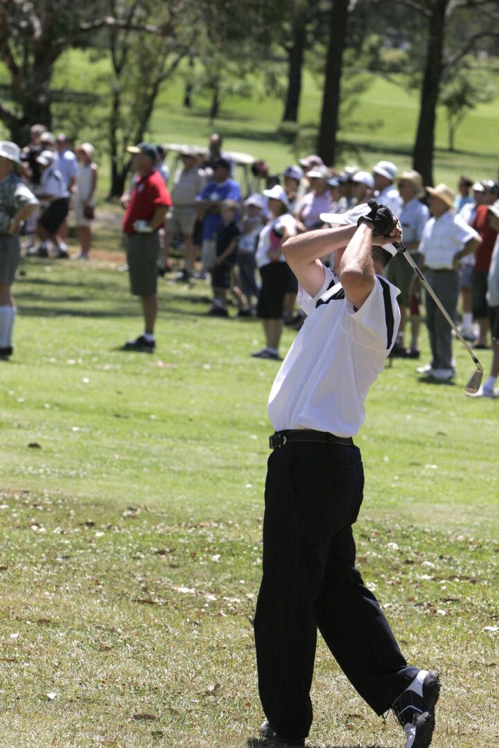 Golfer swinging on course, crowd watching nearby.